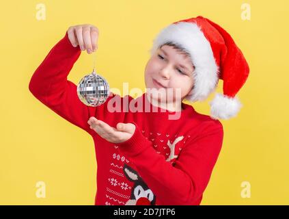 Ein schöner 10-jähriger Junge in einem Weihnachtsmannhut hält eine weihnachtliche Disco-Kugel auf gelbem Hintergrund. Isolieren. Stockfoto