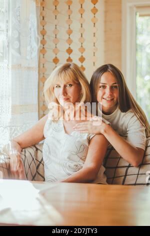 Liebevolle Erwachsene 20s Tochter Umarmung ältere Mutter von hinten während Mutter auf Couch sitzend Menschen posieren Blick auf Kamera lächelnd fühlt sich glücklich, Konzept der mu Stockfoto