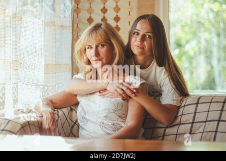 Liebevolle Erwachsene 20s Tochter Umarmung ältere Mutter von hinten während Mutter auf Couch sitzend Menschen posieren Blick auf Kamera lächelnd fühlt sich glücklich, Konzept der mu Stockfoto