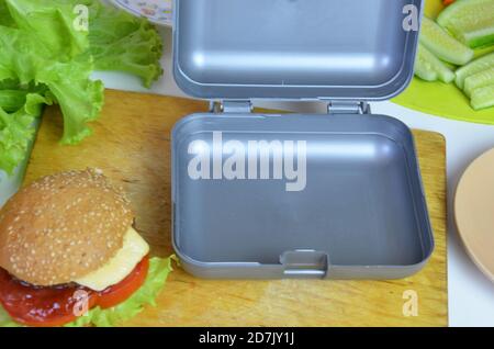 Hausgemachte Hamburger mit Salat in Lunchbox auf dem Tisch. Mit hellgelben Orangensaft. Mit Bananennüssen. Geschnittenes Gemüse Karotten und Gurken Stockfoto