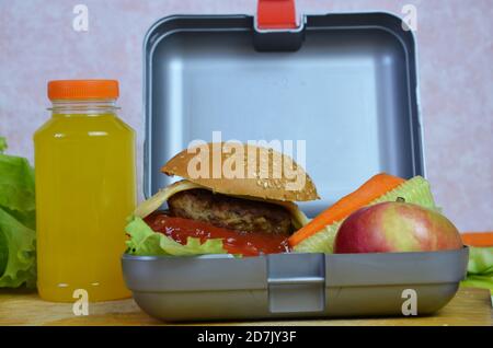 Hausgemachte Hamburger mit Salat in Lunchbox auf dem Tisch. Mit hellgelben Orangensaft. Mit Bananennüssen. Geschnittenes Gemüse Karotten und Gurken Stockfoto