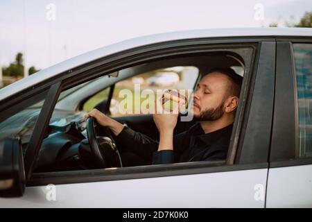 Seitenansicht eines kaukasischen Geschäftsmannes, der einen süßen Donut mit Schokolade in der Hand hält und ein Auto auf dem Weg zur Arbeit fährt. Das Essen eines jungen Mannes Stockfoto