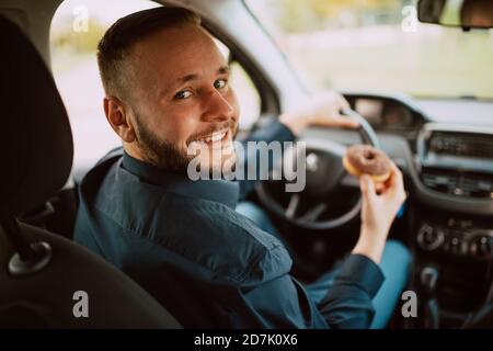 Ein Porträt eines jungen lächelnden kaukasischen Geschäftsmannes, der in einem Auto sitzt und einen köstlichen Schokoladendonut in der Hand hält. Eine süße Mahlzeit beim Transport Stockfoto