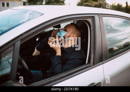 Ein junger Kaukasier, der Gesichtsmaske auszieht, um einen Donut zu essen, während er in einem Auto sitzt, das im Verkehr feststeckt. Schutz gegen COVID - 19 Coronavirus Stockfoto