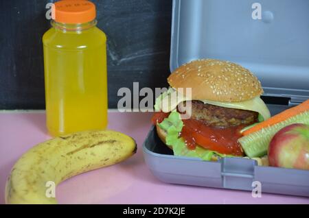 Hausgemachte Hamburger mit Salat in Lunchbox auf dem Tisch. Mit hellgelben Orangensaft. Mit Bananennüssen. Geschnittenes Gemüse Karotten und Gurken Stockfoto