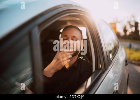 Ein Porträt eines kaukasischen Geschäftsmannes in einem schwarzen Hemd, der in einem Auto sitzt, zur Arbeit geht und einen Schokoladendonut genießt. Süßes Frühstück im Transport Stockfoto