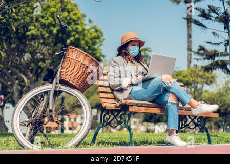 Frau arbeitet in einem Park sitzend und schützt sich mit einer medizinischen Maske. Porträt einer modischen Frau, die die Natur genießt und schreibt auf ihrem Blog Stockfoto