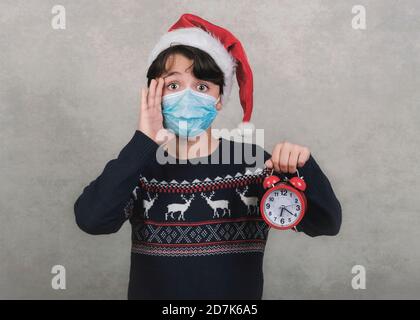 Frohe Weihnachten, Lustiges Kind mit medizinischer Maske und Wecker trägt Weihnachtsmann Hut auf grauem Hintergrund Stockfoto