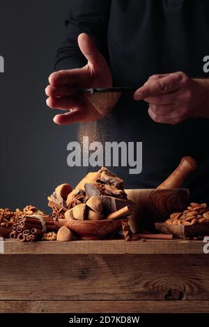 Der Koch streut die Schokolade mit Kakaopulver. Auf einem alten Holztisch Süßigkeiten, Kaffeebohnen, Zimt, Anis, Nüsse, und Stücke von gebrochenem schwarzem Choco Stockfoto