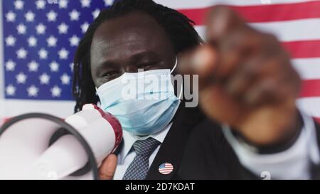 Afrikanischer amerikanischer Mann trägt Gesichtsmaske spricht in Megaphon protestierend Anhebung Klenching Fist - USA Flagge Hintergrund. Hochwertige Fotos Stockfoto