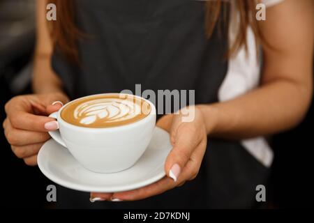 Coffee Business Concept – Nahaufnahme einer Frau, die Kaffee mit Latte-Kunst serviert, während sie im Café steht. Konzentrieren Sie sich auf weibliche Hände, die eine Tasse platzieren Stockfoto