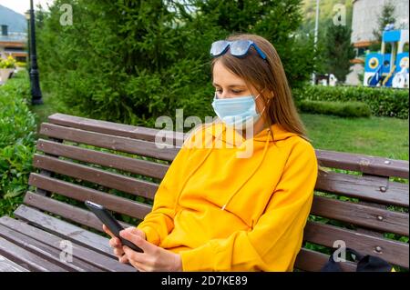 Junge kaukasische Frau in einem gelben Sweatshirt oder Hoodie und Eine Schutzmaske liest ein Smartphone im Park Auf einer Bank Stockfoto