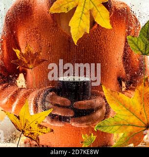 Mann in orange warmer Pullover hält eine Tasse heiße Getränke und schaut durch Fenster mit Herbstblättern. Stockfoto