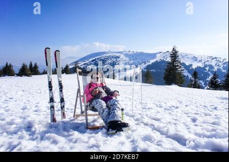 Junge Frau ruht sich aus und trinkt Tee aus einer Thermoskanne nach dem Skifahren auf der Skipiste an einem sonnigen Tag. Lächelndes Mädchen mit Skifahren. Stockfoto