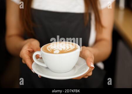 Coffee Business Concept – Nahaufnahme einer Frau, die Kaffee mit Latte-Kunst serviert, während sie im Café steht. Konzentrieren Sie sich auf weibliche Hände, die eine Tasse platzieren Stockfoto