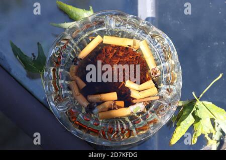 Ein Glas Aschenbecher auf einem Glastisch, es wird verwendet und es hat etwas Wasser in ihm. Der Tisch hat auch ein paar Blätter darauf. Stockfoto