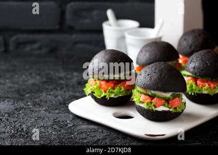 Schwarzer Burger mit geräuchertem Lachstartar, Gurke, Salat und Frischkäse, serviert auf einer weißen Keramikplatte, schwarzer Hintergrund, selektiver Fokus Stockfoto