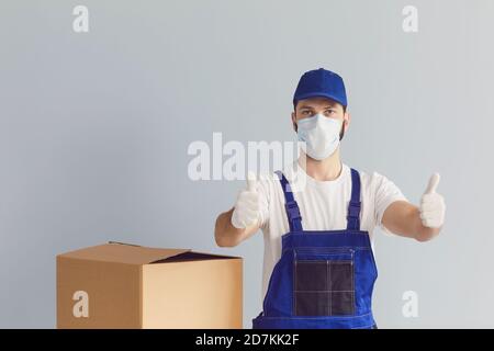 Männlicher Kurier in Uniform, medizinische Maske und Handschuhe hebt Daumen hoch auf grauem Wandhintergrund. Stockfoto