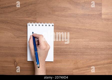 Hand einer jungen kaukasischen Frau, die mit einem blauen Stift auf einer leeren Seite eines offenen Notizbuchs auf einem Holzschreibtisch schreibt. Bild von oben mit Kopierbereich. Stockfoto