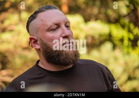 Hipster Stil bärtiger Mann. Der attraktive Mann die Blondine mit langen Haaren des europäischen Auftritts mit einem Bart. Mann in braunem T-Shirt. Mann im Wald Stockfoto