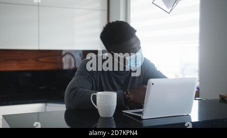 Junger afroamerikanischer schwarzer Mann, der in seiner Wohnung am Laptop arbeitet, während er Gesichtsmaske trägt. Hochwertige Fotos Stockfoto