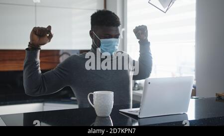 Junger afroamerikanischer schwarzer Mann, der sich beim Arbeiten am Laptop in seiner Wohnung mit Gesichtsmaske ausdehnt. Hochwertige Fotos Stockfoto