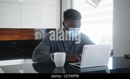 Junger afroamerikanischer schwarzer Mann, der in seiner Wohnung am Laptop arbeitet, während er Gesichtsmaske trägt. Hochwertige Fotos Stockfoto