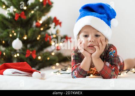 frohe Weihnachten und glückliches neues Jahr, Kindheit, Urlaub Konzept - close-up kleine traurige Mädchen in blau weihnachtsmann Hut auf seinem Bauch spielen und viel Spaß auf dem Bett Stockfoto