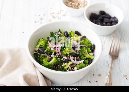 Brokkoli mit Rosinen, roten Zwiebeln und Samen. Gesunde Rohkost Salat. In einer weißen Schale auf blauem Holzhintergrund Stockfoto