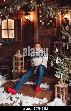 Ein hübscher junger Mann in warmer Kleidung sitzt darauf Die Stufen im Hof des bepflanzten Hauses davor Das neue Jahr Stockfoto
