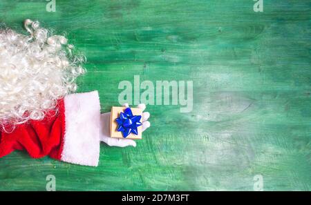 Draufsicht auf eine Weihnachtsmann-Handschaufel zu Weihnachten mit einem kleinen Geschenk mit blauer Schleife auf grünem Holzhintergrund sieht man ein Detail des Bartes Stockfoto