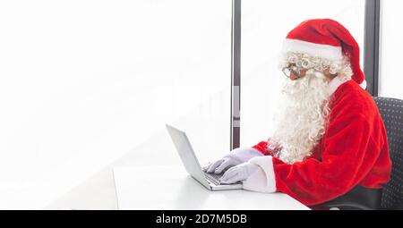 Weihnachtsmann arbeitet an einem Laptop in einem minimalistischen weißen Büro zu Weihnachten. Textbereich Stockfoto