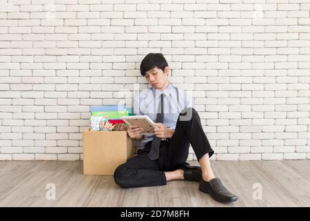 Ein asiatischer Mann saß traurig auf einem Holzboden in einem Büro, nachdem er gefeuert wurde, und speicherte persönliche Gegenstände in Kartons. Stockfoto