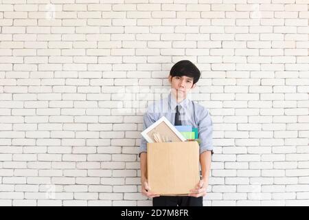 Ein asiatischer Mann steht traurig in einem Büro, nachdem er gefeuert wurde, und hält persönliche Sachen in Kartons. Stockfoto