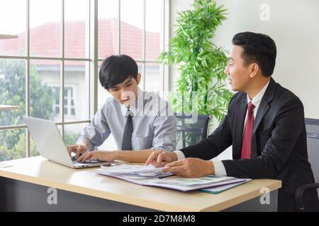 Ein asiatischer männlicher Supervisor bringt dem Neuling auf seinem Schreibtisch das Büro bei. Stockfoto