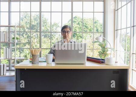 Asiatischer Mann mittleren Alters, der an einem Schreibtisch in einem Büro sitzt. Stockfoto