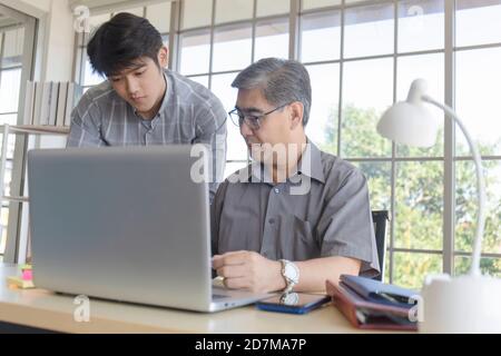 Ein asiatischer Mann mittleren Alters, der seinem Sohn einen Job beibringt, der neben einem Schreibtisch in einem Büro steht. Stockfoto