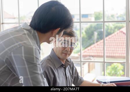 Ein asiatischer Mann mittleren Alters, der seinem Sohn einen Job beibringt, der neben einem Schreibtisch in einem Büro steht. Stockfoto