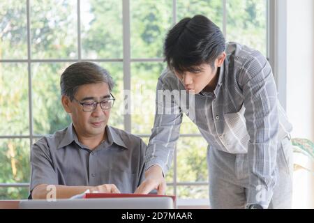 Ein asiatischer Mann mittleren Alters mit seinem Sohn, der neben einem Schreibtisch im Büro steht. Stockfoto