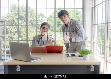 Ein asiatischer Mann mittleren Alters mit seinem Sohn, der neben einem Schreibtisch im Büro steht. Stockfoto