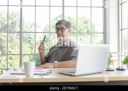 Ein asiatischer Mann mittleren Alters, der einen Stift hält und darüber nachdenkt, Probleme auf einem Schreibtisch in einem Büro zu lösen. Stockfoto