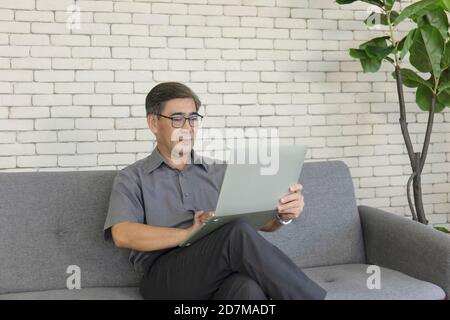 Ein asiatischer Mann mittleren Alters, der ein Notebook in der Hand hält, sitzt auf einem Sofa und nimmt einen Videoanruf ab. Stockfoto