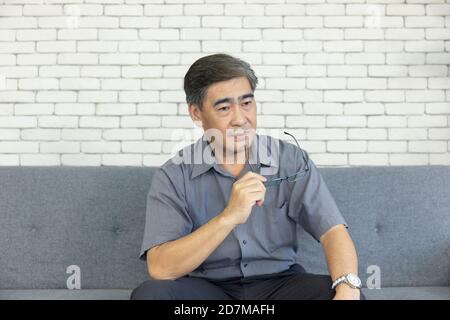 Ein asiatischer Mann mittleren Alters in seiner Handbrille und war wie das Denken auf dem Sofa sitzen. Stockfoto