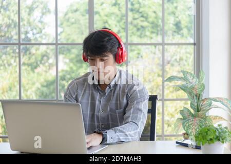 Ein junger asiatischer Geschäftsmann arbeitet mit Kopfhörern auf einem Balkon-Schreibtisch an einem Glasfenster. Stockfoto