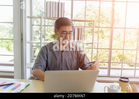 Ein asiatischer Mann mittleren Alters verwendet seine Kreditkarte, um online über einen Laptop auf seinem Schreibtisch einzukaufen. Stockfoto