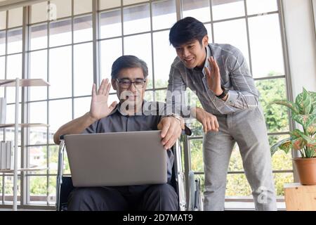 Ein asiatischer Vater mittleren Alters sitzt in einem Rollstuhl und hat einen Sohn neben ihm, der bei Videoanrufen auf einem Laptop chattet. Stockfoto