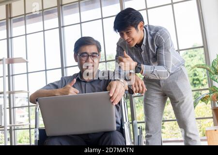 Ein asiatischer Vater mittleren Alters sitzt in einem Rollstuhl und hat einen Sohn neben ihm, der bei Videoanrufen auf einem Laptop chattet. Stockfoto