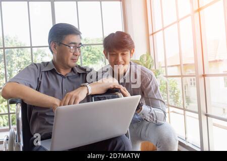 Ein asiatischer Vater mittleren Alters sitzt in einem Rollstuhl und hat einen Sohn neben ihm, der bei Videoanrufen auf einem Laptop chattet. Stockfoto