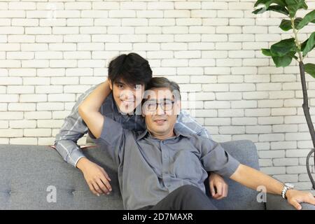 Ein Vater mittleren Alters und ein asiatischer Sohn umarmten sich auf dem Sofa mit einem glücklichen Lächeln. Stockfoto