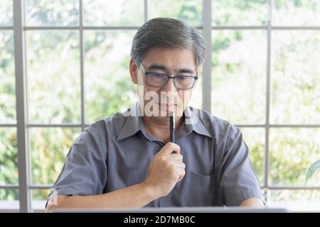 Ein älterer asiatischer Mann, der eine denkende Geste in seinem macht Hand hält einen Stift Stockfoto